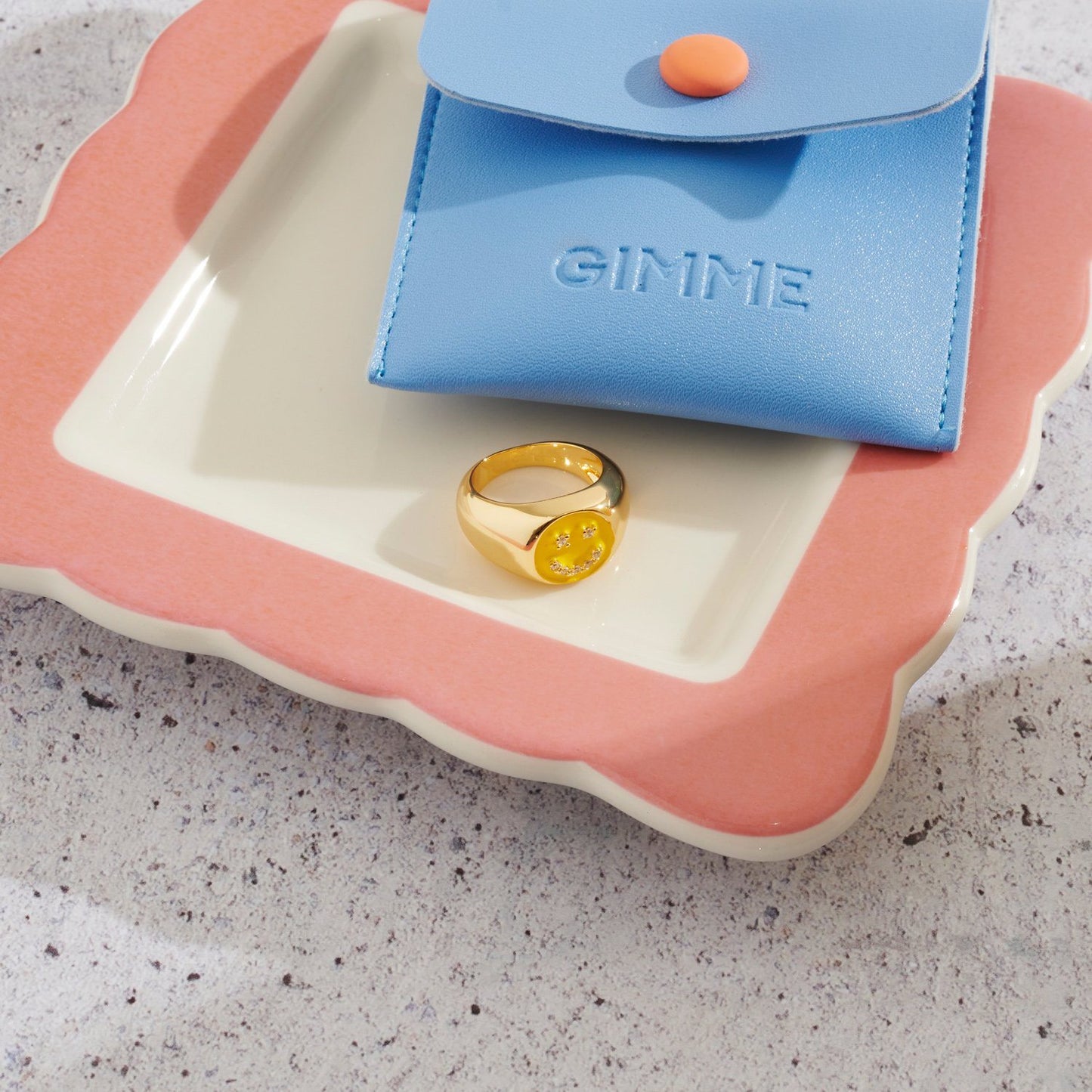 Chunky ring with yellow smiley face sitting on a plate next to jewellery pouch.